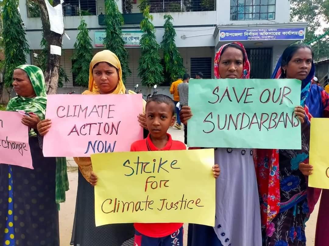 Several South Asian women in colorful outfits and a young boy stand on a street in Bangladesh. They have serious facial expressions, and they are holding paper signs that say "Climate Action Now," "Strike for Climate Justice," and "Save Our Sundurbans."