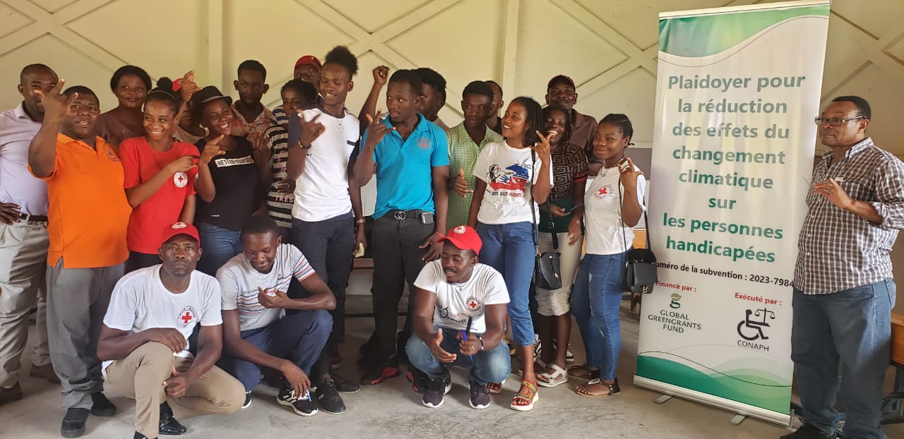 A group of people from Haiti stand together as a group and pose for the camera. They are all Black and have mixed gender presentations. Most of them are holding their hand in one position, with their thumb, pointer, and pinky finger up. Many of them are laughing. Next to them is a banner that says in French: "Plaidoyer pour la reduction des effets du changement climatique sur les personnes handicapees." Beneath that text are logos for Global Greengrants Fund and our Haitian grantee partner CONAPH.