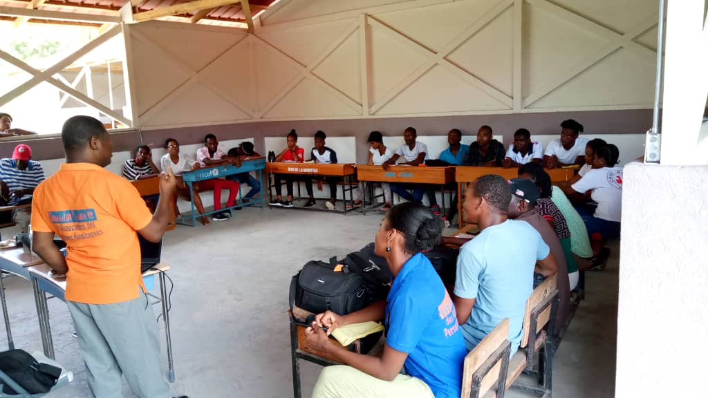 Emilio Neas's back faces the camera. He is wearing an orange t-eeshirt with the Coalition Nationale des Personnes Handicapées’ logo. He is talking to a room of 13 people, who are all sitting down and looking at him.