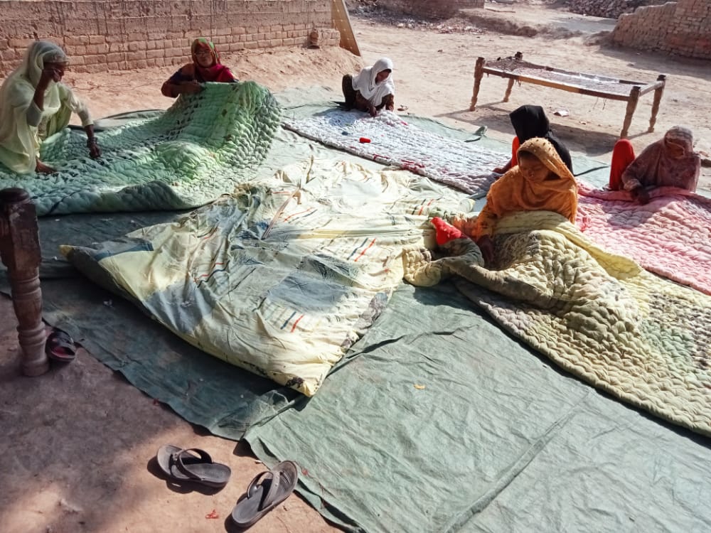Six women sit among blankets that are spread out over the ground. They are in the midst of creating the blankets.