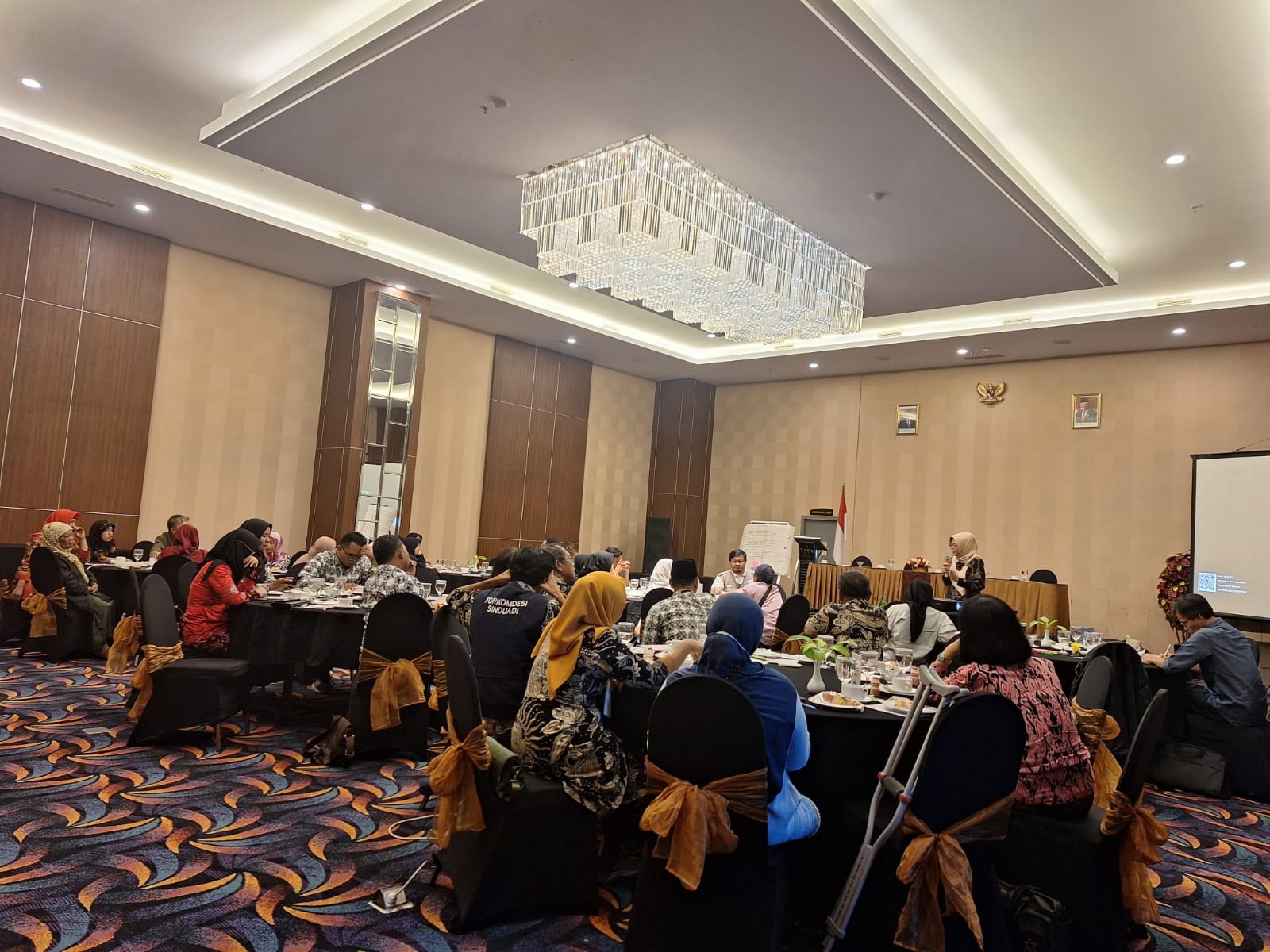 A zoomed out view of the conference room where a Sinduadi workshop was held. Participants are seated around round tables while a speaker stands at the front of a room with a microphone.