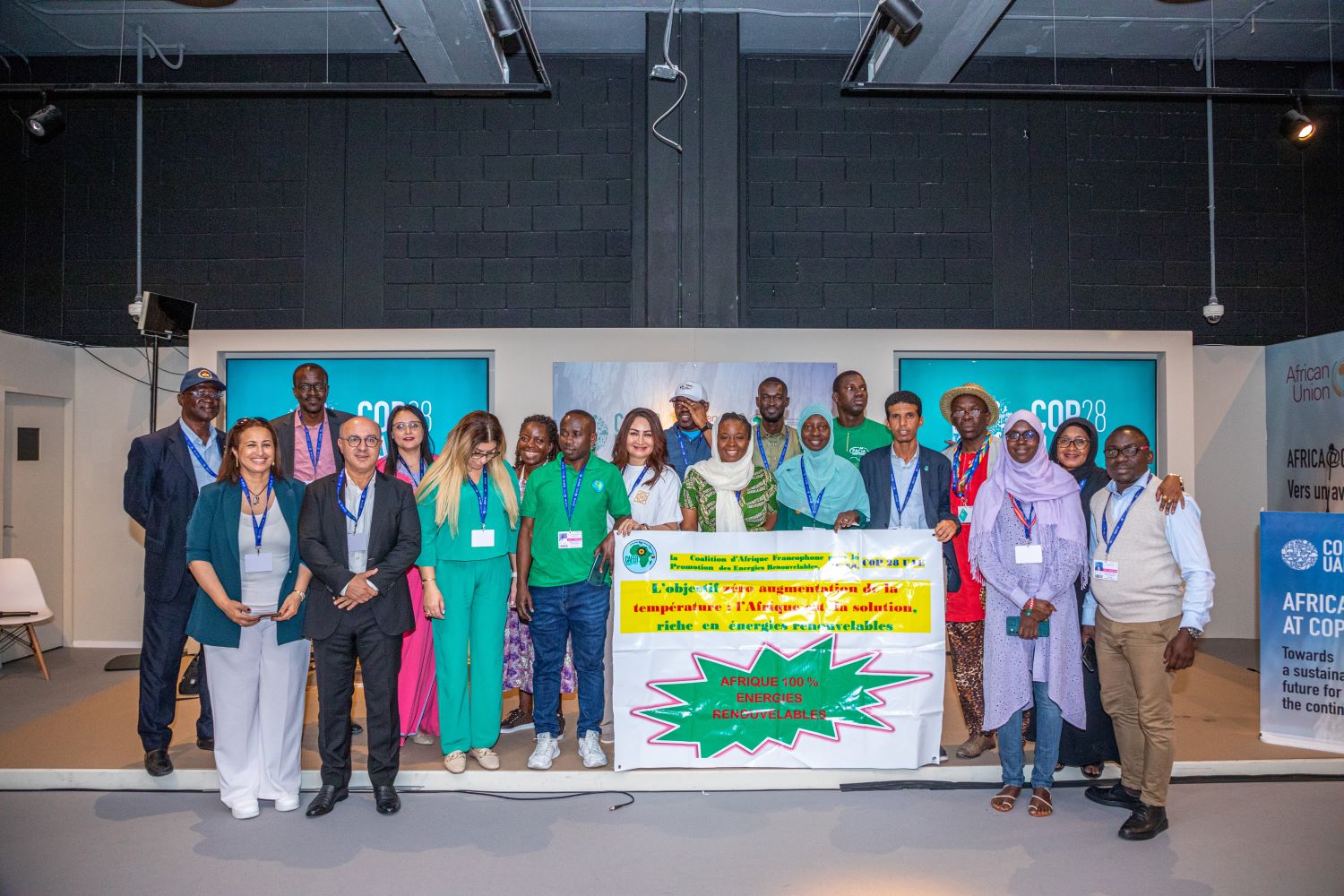 A group of people of varying races and gender presentations stand together on stage around a banner with text in French. Behind them are screens that say "COP28."