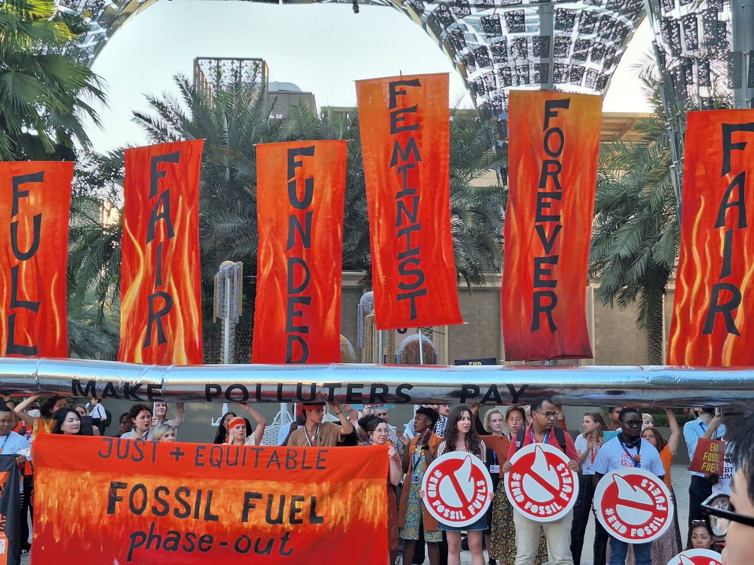 A group of people of varying races and gender presentations stand together at an outdoor protest. They are holding banners with firing background colors that read "Full, Fair, Funded, Feminist, and Forever" as well as "Just + Equitable Fossil Fuel Phase-Out."