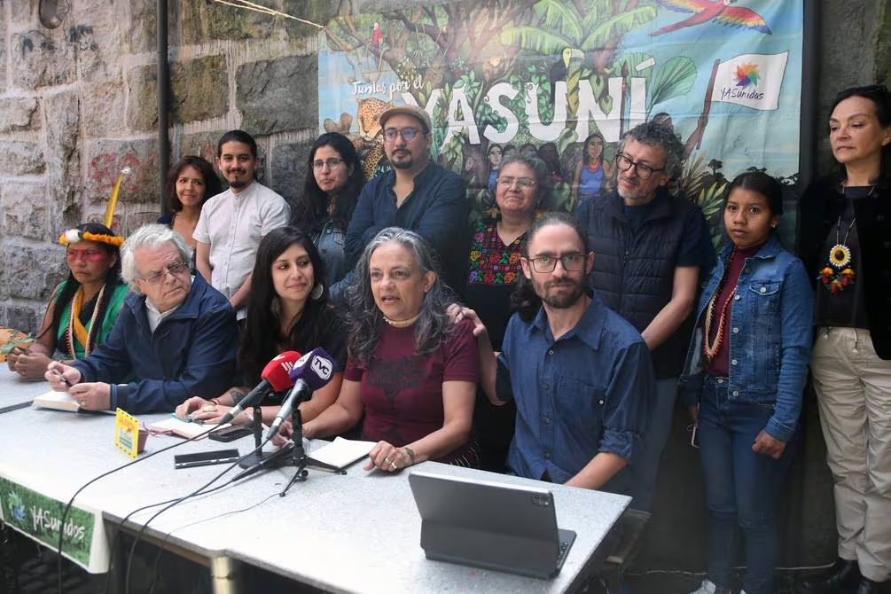 13 Latine people of varying ages and gender presentations gather around a table, presumably facing an audience. Some are seated at the table, and some are standing in a line behind it. One person seated at the table is speaking into a microphone, while many of the others are looking at them with slight smiles. On the wall in the background is a banner with colorful imagery of trees and animals, with bold text that says "Yasuni."