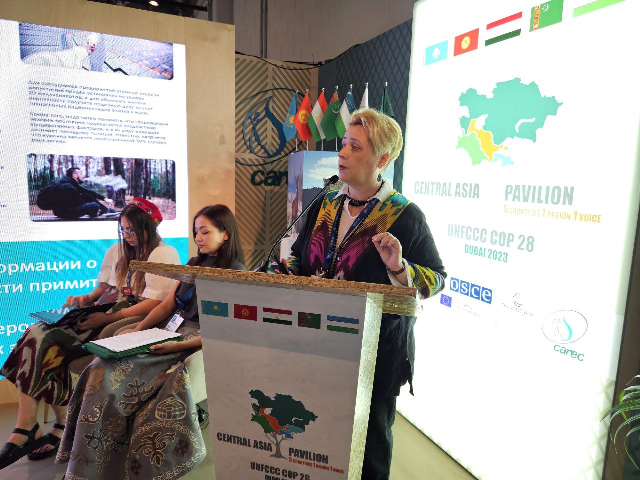 A woman with short blonde hair stands at a podium, in the middle of speaking. The podium has Central Asia flags on it and a COP28 logo, and says "Central Asia Pavilion." Next to her, two women are seated with notes in their laps. Behind them is a screen with a map of Central Asia, Central Asia flags, and the text "Central Asia Pavilion."