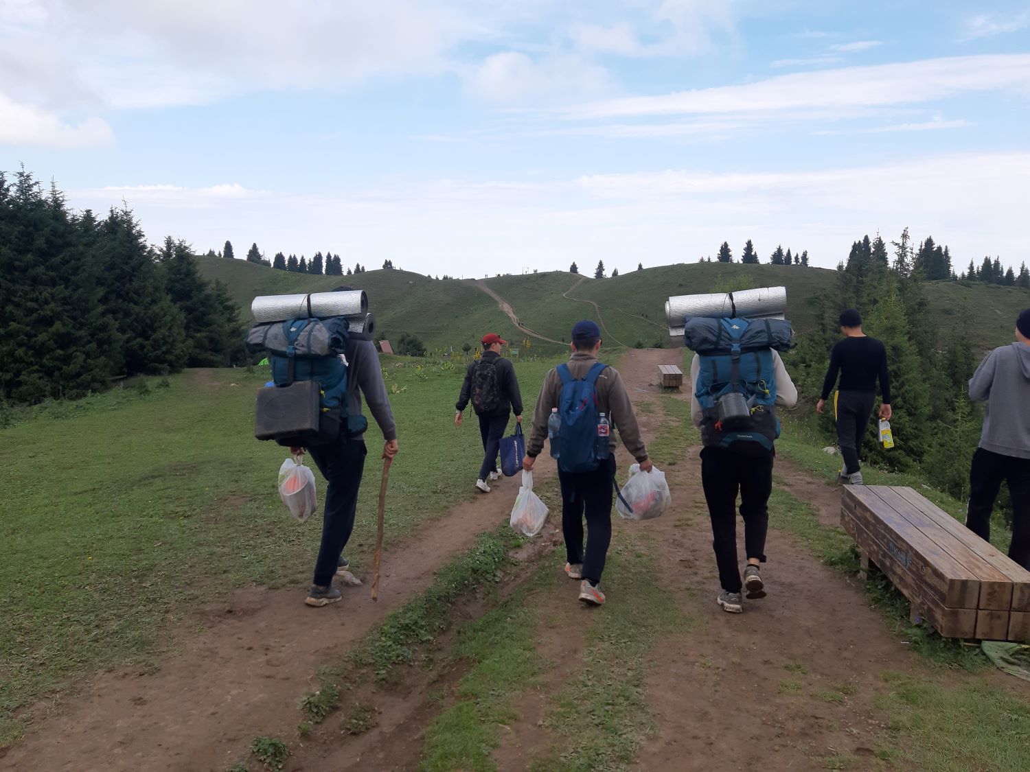 Six people hike uphill on a dirt trail that extends off into the distance. They are facing away from the camera. Around them are grass and a few trees, and lightly cloudy skies. Many of the people are carrying large backpacks, and some are carrying small plastic bags filled with trash.