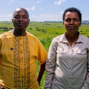 Sinegugu and Nonhle stand next to each other. Sinegugu is on the left. He is a Black man with a bald head. He is wearing a bright yellow shirt with colorful patterns embroidered onto it, and he is smiling slightly, his hands on his hips. On the right is Nonhle. She is a Black woman with curly hair cropped short. She is wearing a button up grey shirt and a more serious expression. Behind them is a lush and bright green field of grass.