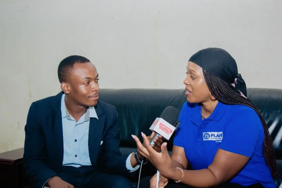 Two Black people sit together on a couch, facing each other. On the left, a Black man holds a microphone up to the Black woman on the right. He is looking at her with an engaged expression, while she is in the middle of speaking.