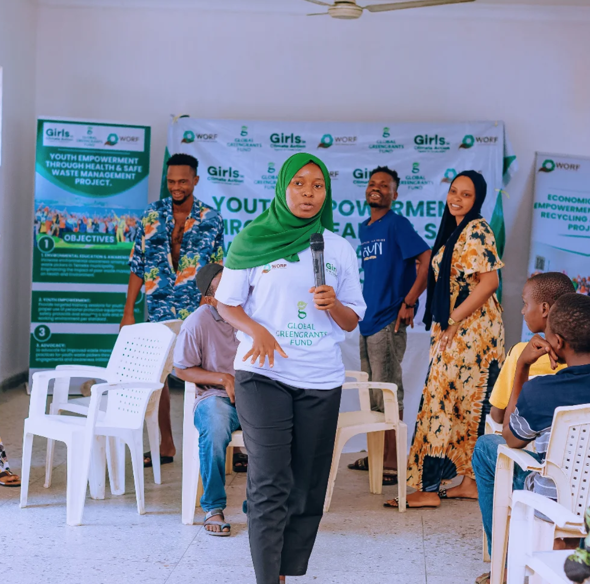 A Black woman with a headscarf stands in the center of a room with a microphone, in the middle of speaking. In the background, workshop participants stand and sit, many looking at the woman with the microphone, and many smiling. In the background are banners that read "Youth Empowerment through Health & Safe Waste Management Project."
