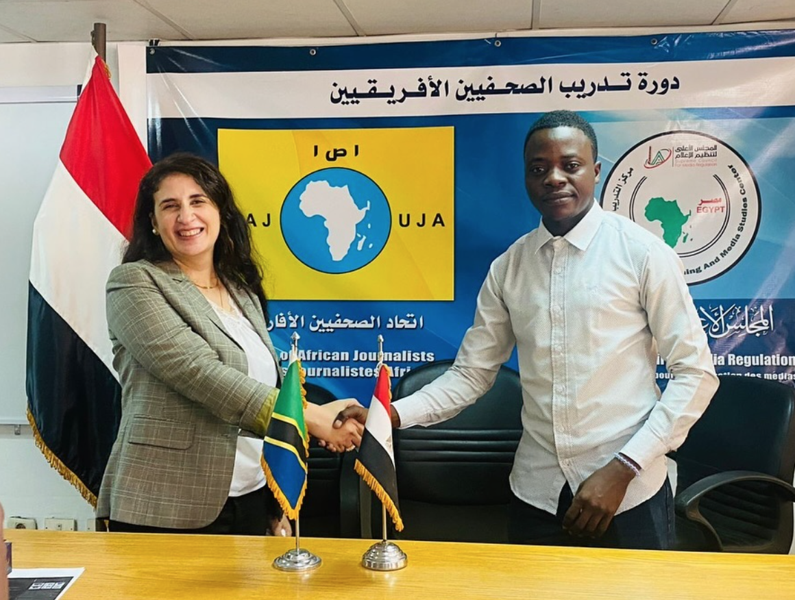 Two people, a brown-skinned woman and Black man, pose together for the camera, shaking hands. On the table in front of them are Egyptian and Tanzanian flags.