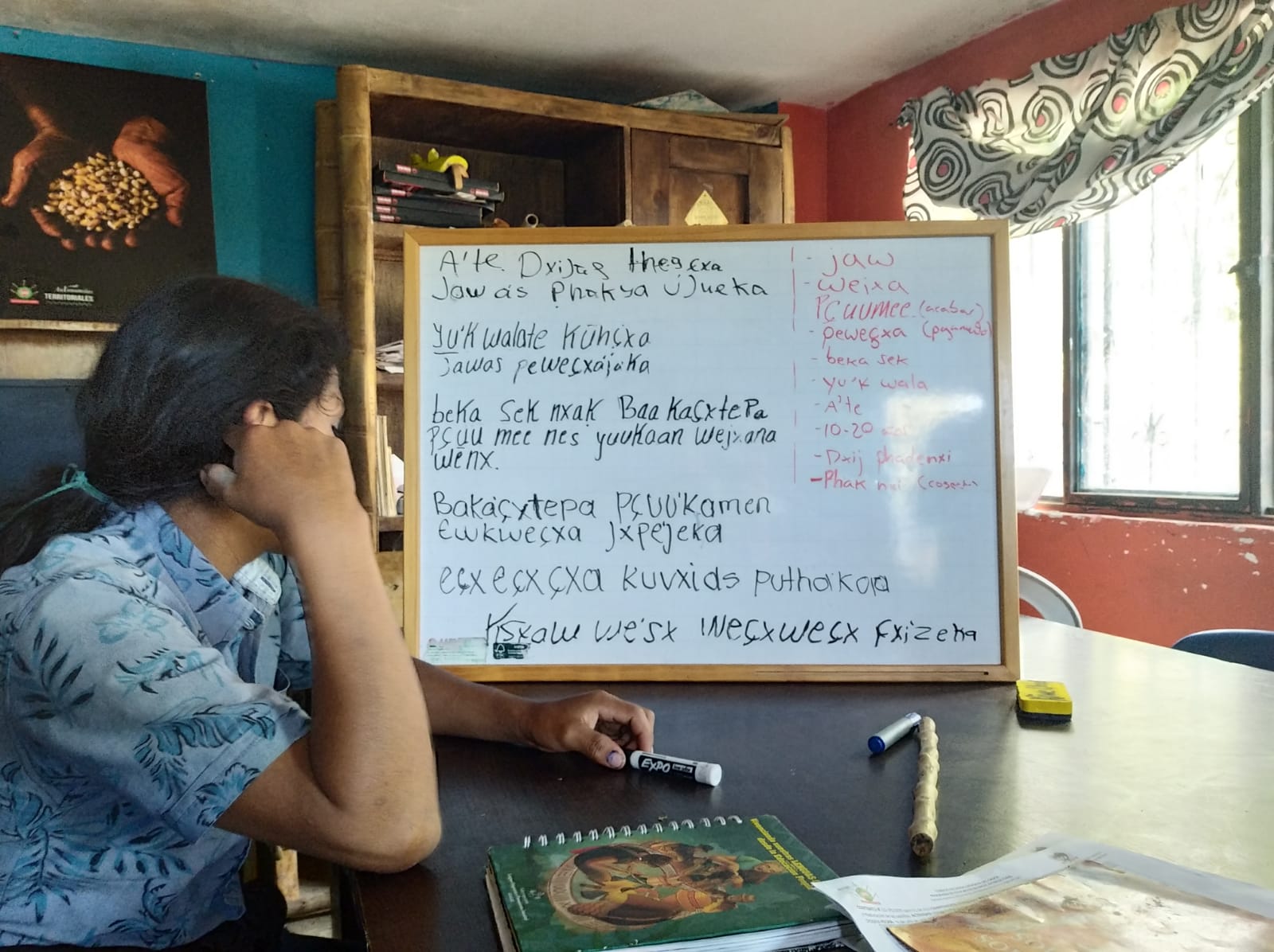 A Latine young person sits at a table holding a marker. On the table is a white board with text in an Indigenous language.