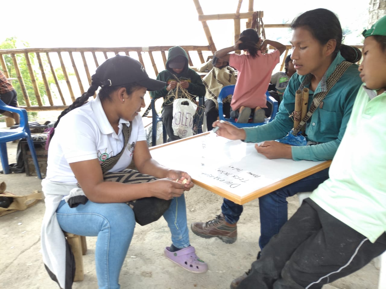 Three Latine youth sit together around a white board. Two of them seem to be in the middle of speaking, and one has their hand poised with a marker over the board.