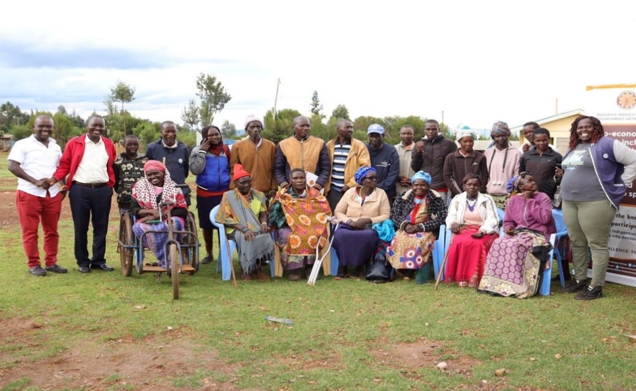 A group of nearly two dozen Black people of varied gender presentations poses together for a group photo, many of them smiling. Some of them have visibility disabilities, for instance one is sitting in a wheelchair and one has a crutch leaning against their chair.