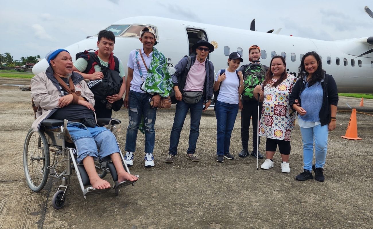 A group of people with varying gender presentations and disabilities, visible and not, pose together in the Philippines. Many of them are smiling. Behind them is a small plane.