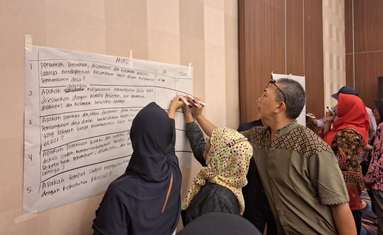 Three people with varying gender presentations face away from the camera and write on a large piece of paper that is taped to a wall. The text on the page is in Indonesian.