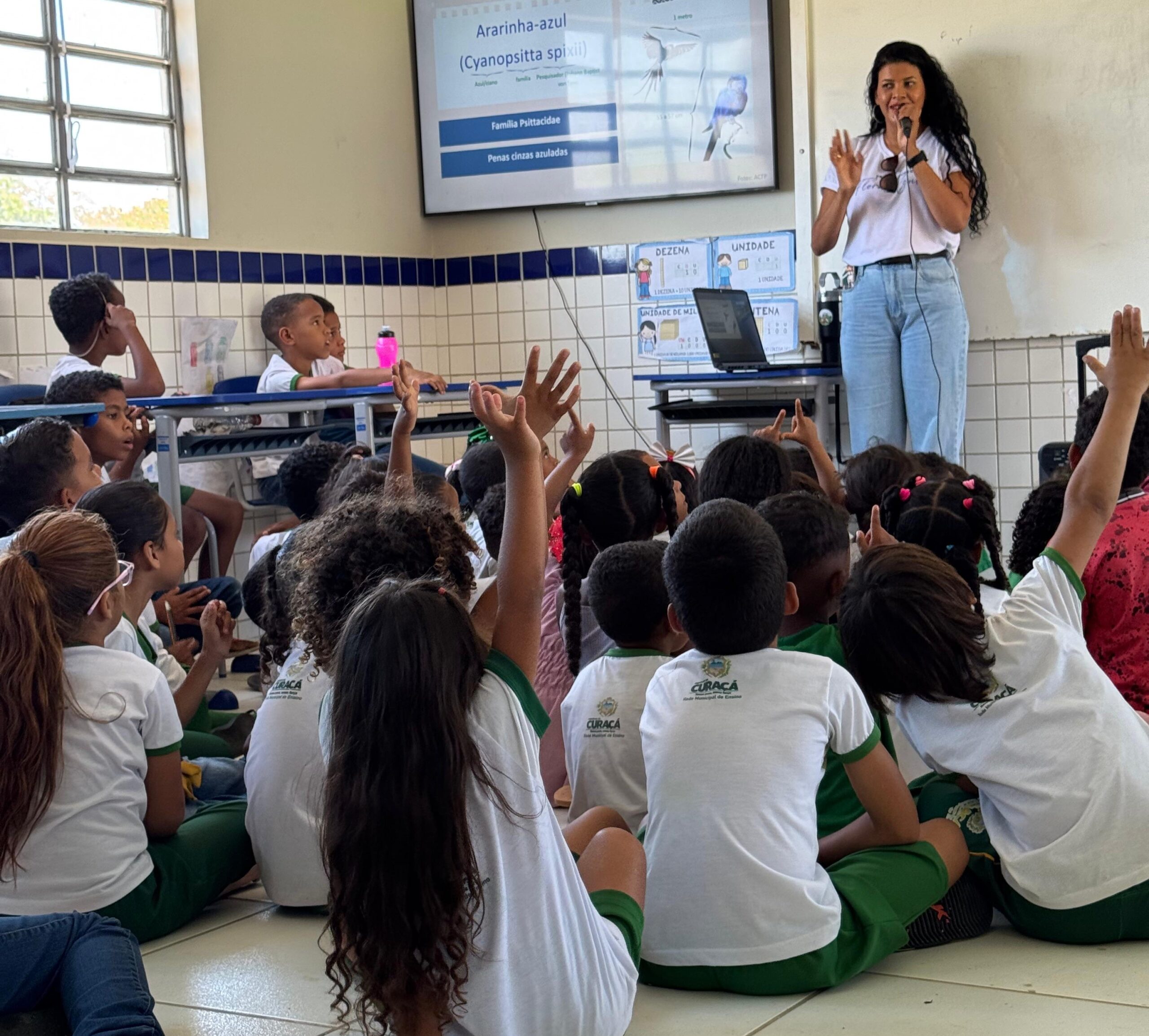 A Latine woman stands in front of a group of children, many of whom are raising a hand. The woman is holding a microphone, in the middle of speaking.