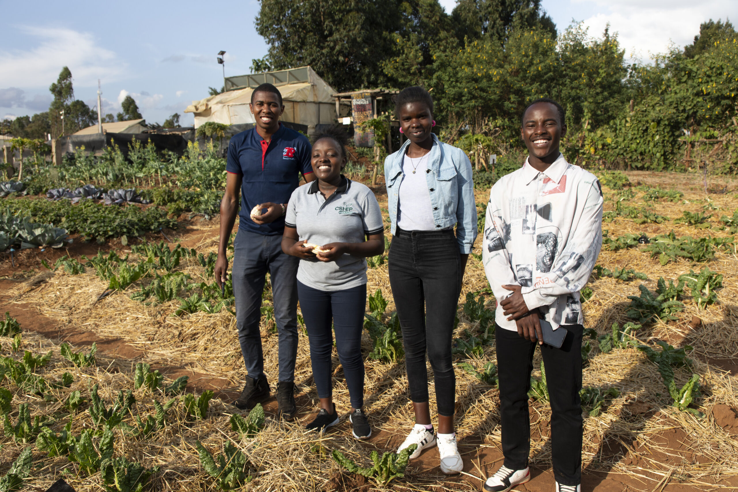 Four Black youth with mixed gender presentations stand together in a garden. They are looking at the camera and smiling brightly.