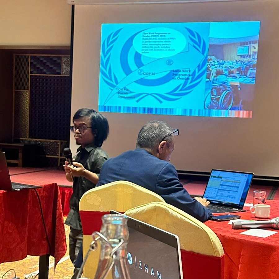 An Asian little person stands in front of a projector screen on which is displayed a presentation slide. They are holding a microphone, in the middle of speaking.