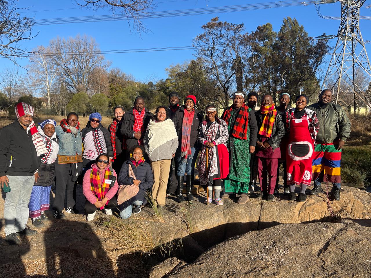 Over a dozen people of varied gender presentations and races stand together posing for a photo, many of them smiling. Many of them are wearing colorful Indigenous accessories are articles of clothing.
