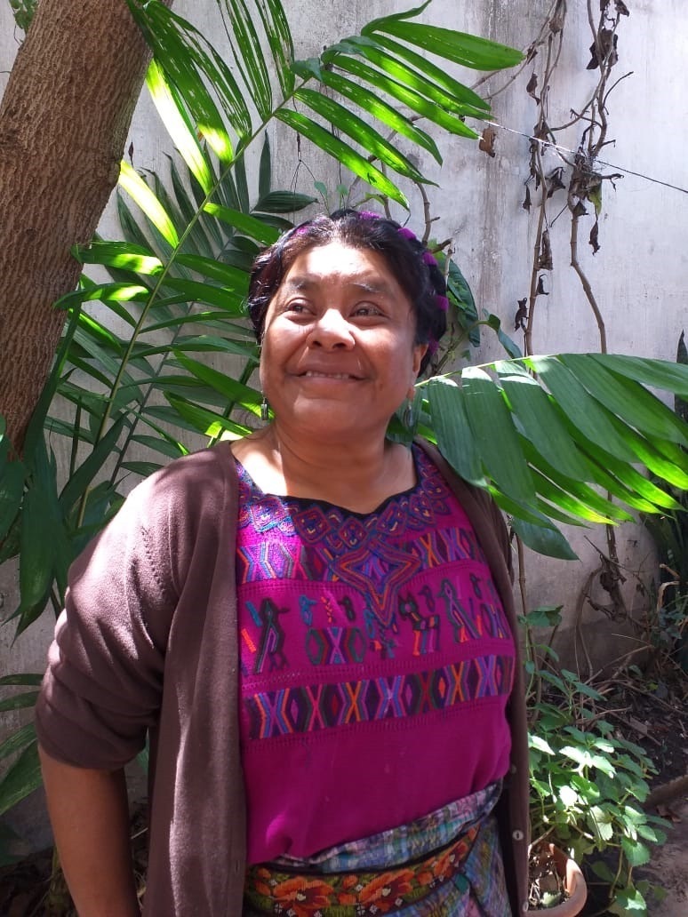 A photo of María Huz. She is a brown-skinned woman wearing a purple shirt with colorful patterns on it and a sweater. She is looking off to the left and smiling warmly. Behind her is a tree.