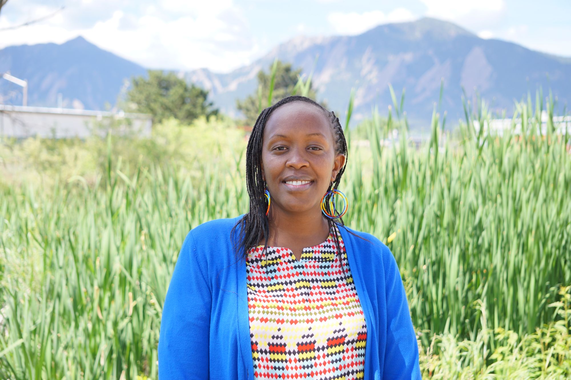 A photo of Naomi Leleto. She is a Black woman with braids, colorful circle-shaped earrings, and a colorfully-patterned shirt with a bright blue sweater. She is looking at the camera and smiling. Behind her is a field of tall grass, and behind the grass are mountains.
