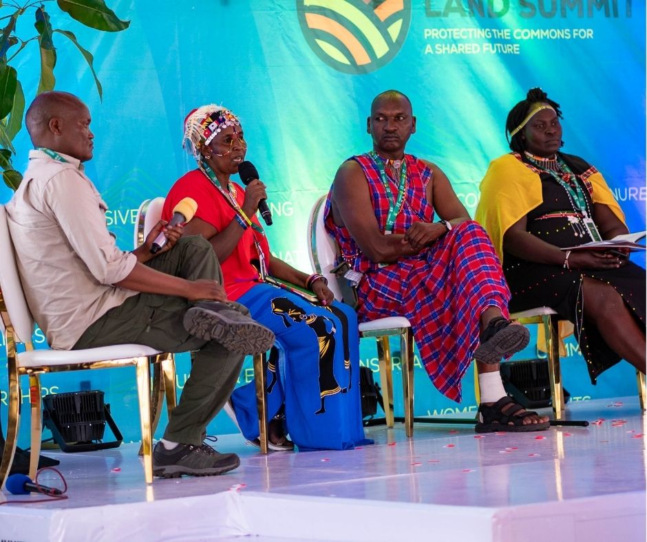 Four Black people, many of them wearing elements of Indigenous clothing and accessories, sit together on a stage. One person is holding a microphone and in the middle of speaking. Behind them is a blue banner that says "Land Summit" Protecting the Commons for a Shared Future."
