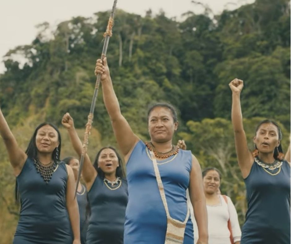 Several Indigenous women from Ecuador stand together smiling with fists raised. A woman in the foreground is holding up a spear. Most of them are wearing blue dresses and colorful beaded necklaces.