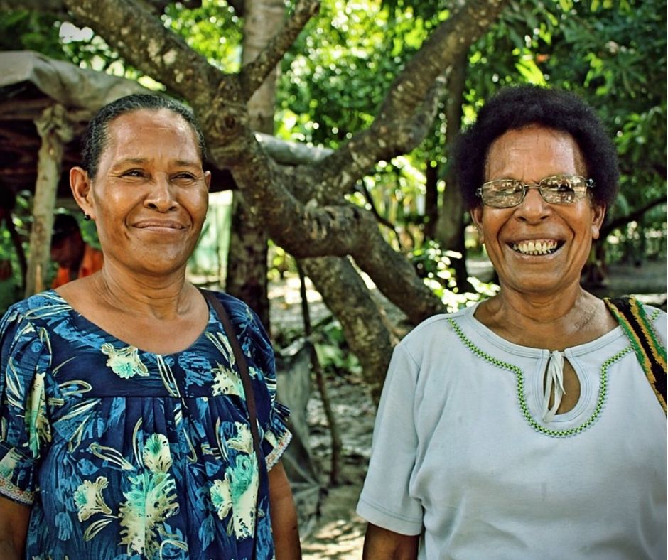 Two older Brown women pose together for the camera, both smiling warmly. Behind them is a tree and a lot of greenery.