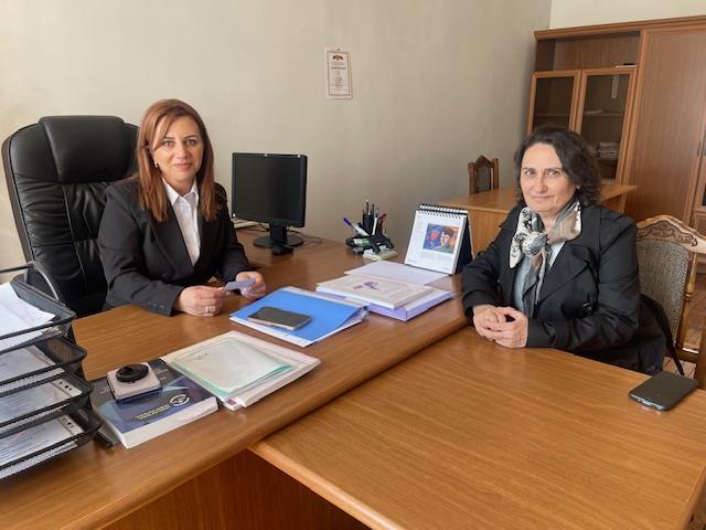 Two women in formal clothes sit together at desks, smiling slightly at the camera.