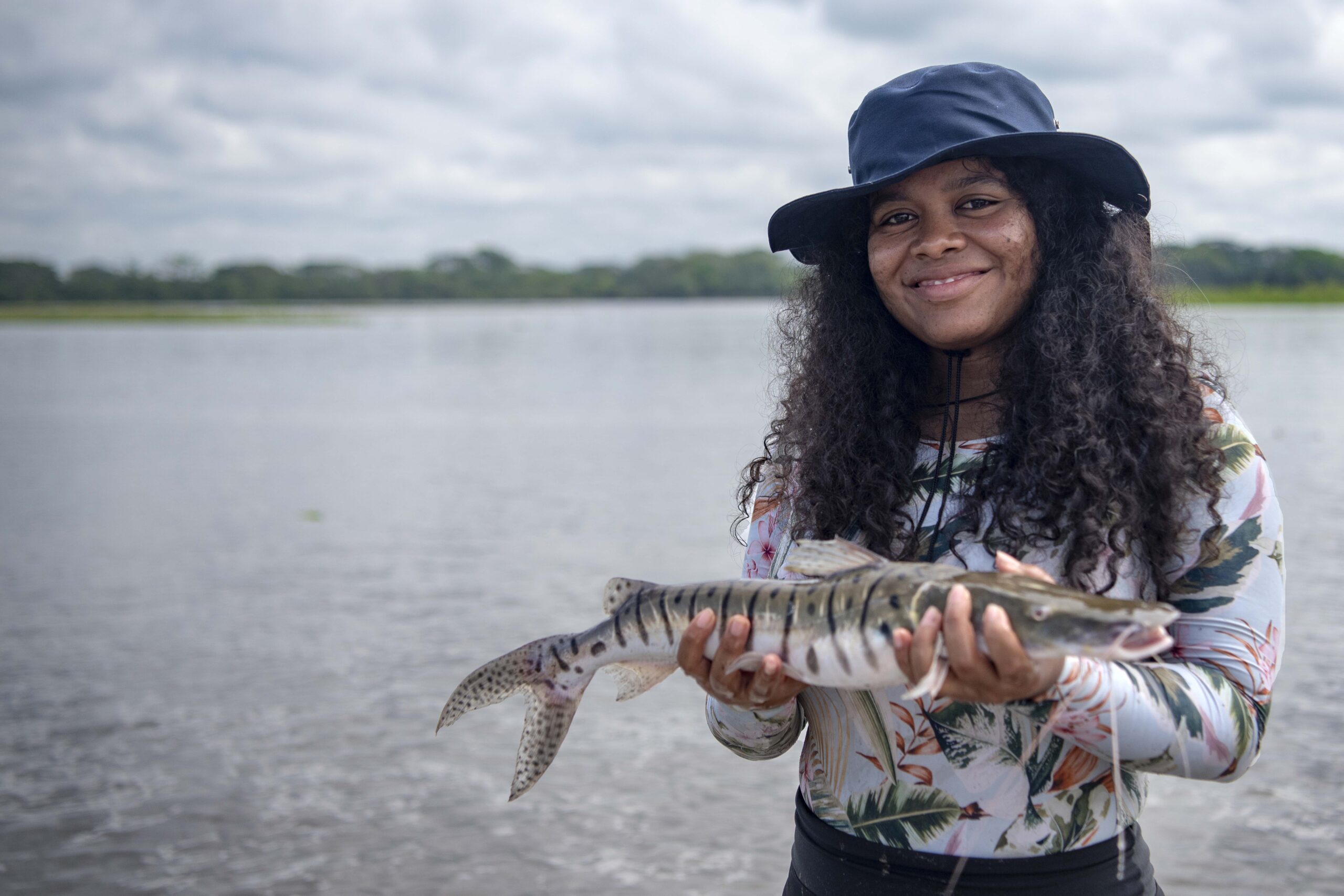 A portrait of Yuvelis Morales Blanco. She is looking directly at the camera and smiling, holding a fish. She is a Brown woman with long dark curly hair, and she is wearing a floral print shirt and a dark blue fishing hat. In the background is the Magdelena River.