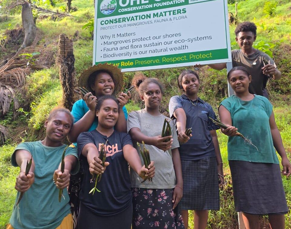 A group of Pacific Islander youth pose together for the camera, smiling and holding up mangrove seedlings. Behind them is a sign with the title "Conservation Matters" in bold font.