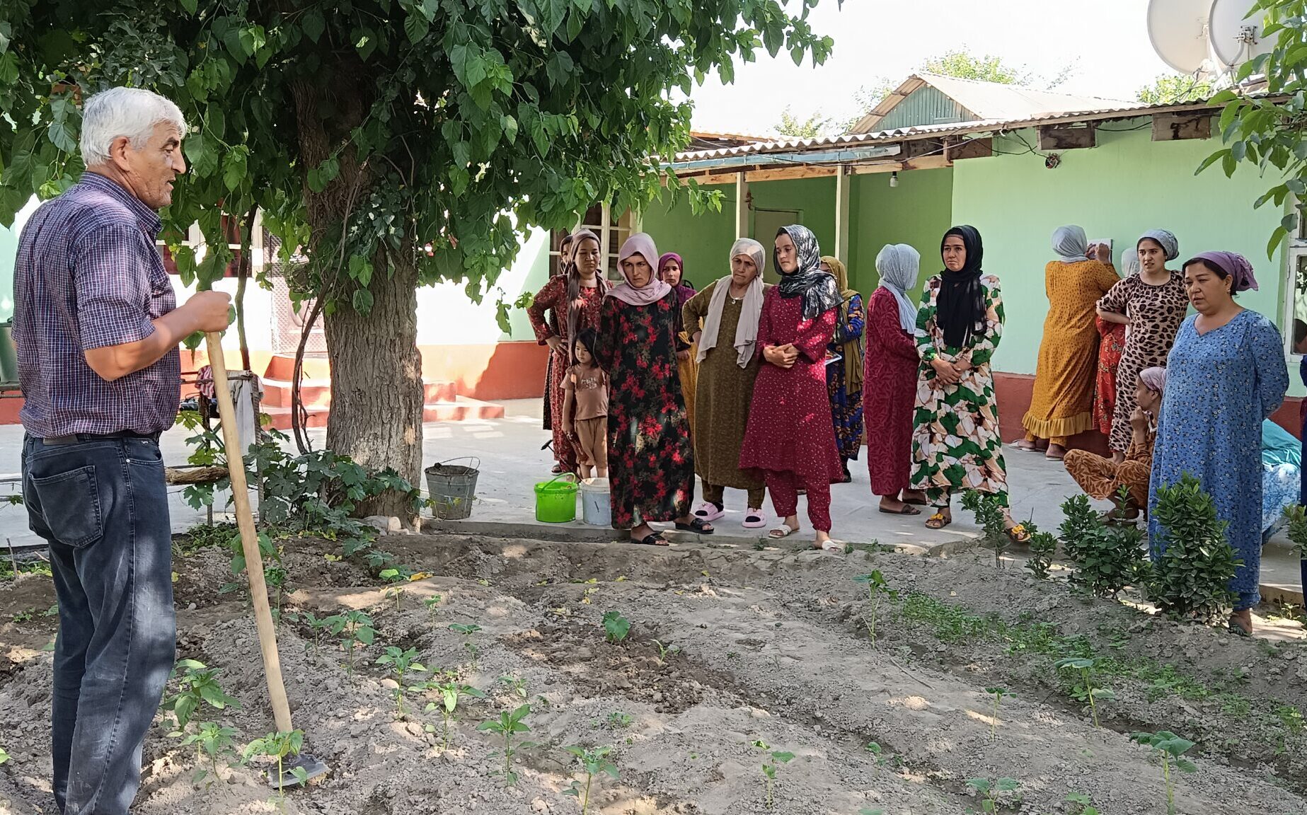 On the left side of the photo, a man stands in the middle of a garden with a long gardening tool, in the middle of speaking. On the right, at the edges of the garden, women in colorful Tajik clothing are looking at the man, seemingly listening intently.