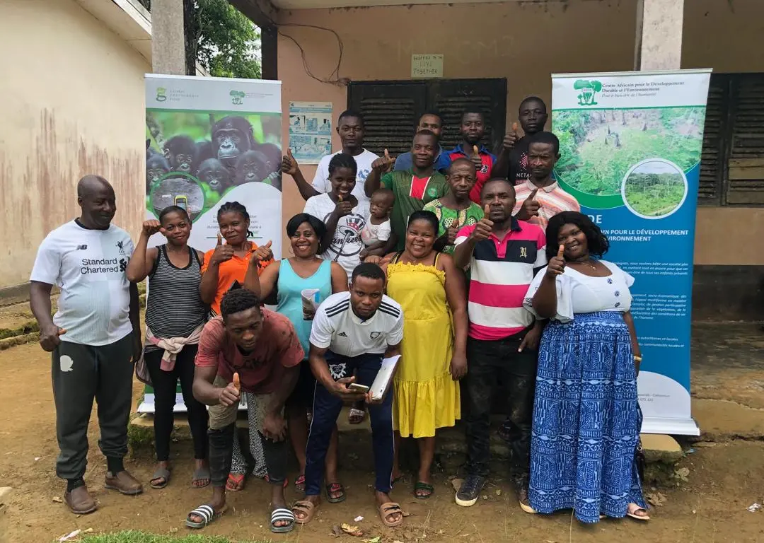 A group of Black people with varied gender presentations pose together for the camera. Many of them are smiling and holding up a thumbs up sign. Behind them are signs featuring photos of gorillas and forests.