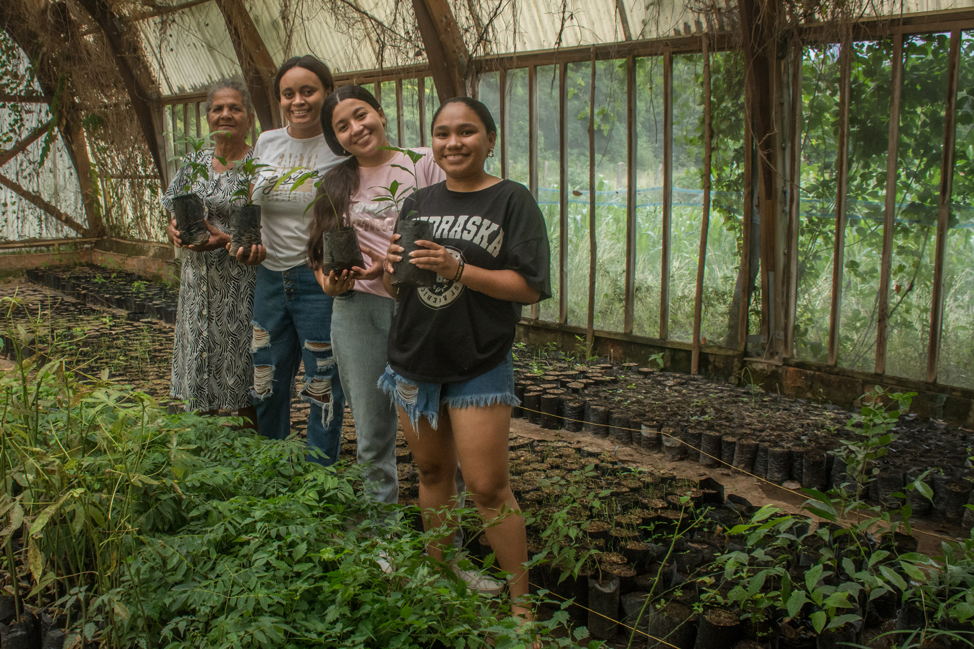 Four Latina women of mixed ages stand together in a greenhouse, each holding a young plant. They are all smiling.