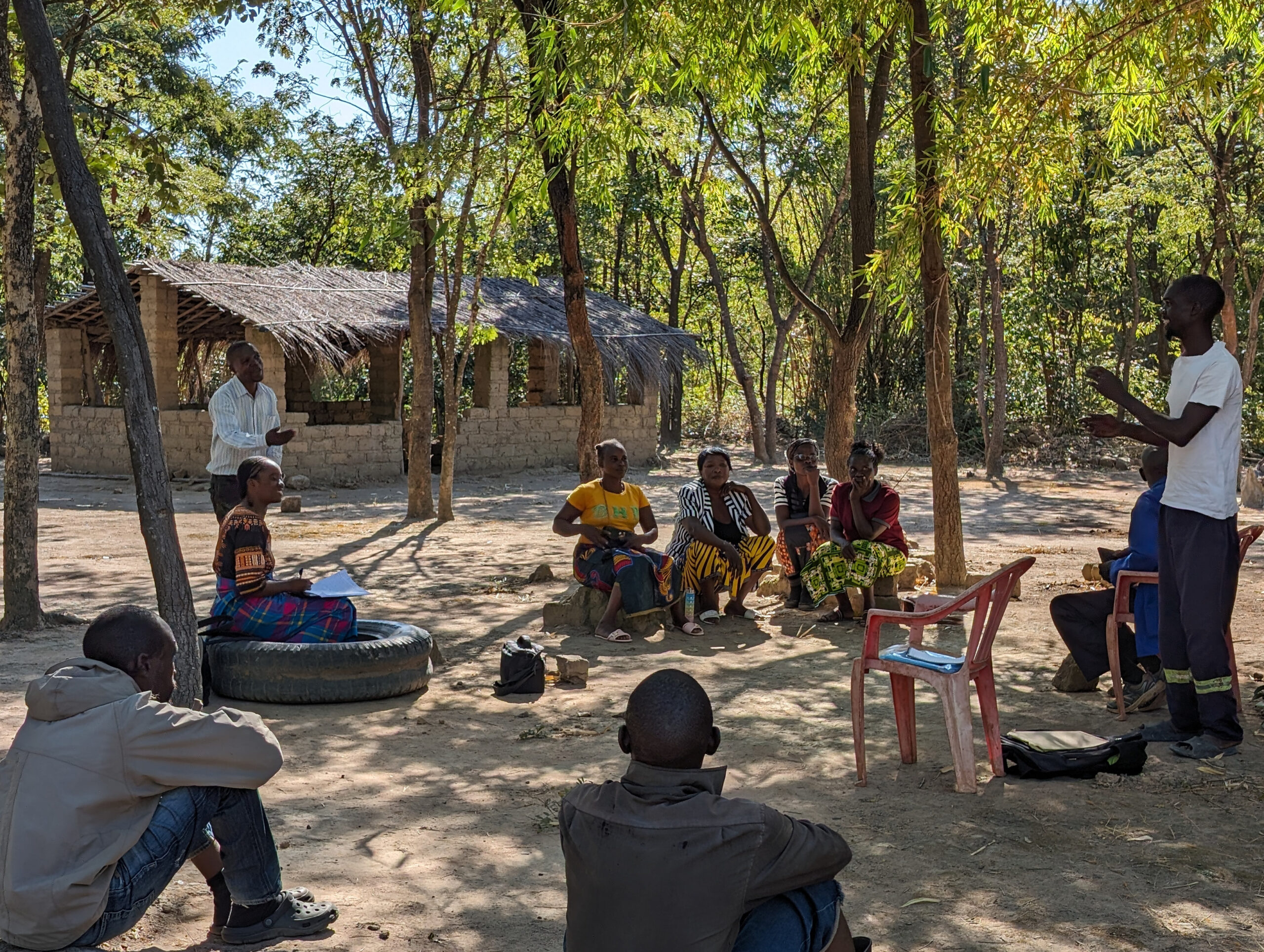 A group of Black people with mixed gender presentations sitting in a circle outside. To the left and right, two men are standing and gesturing as if in the middle of speaking. Around them are many trees.
