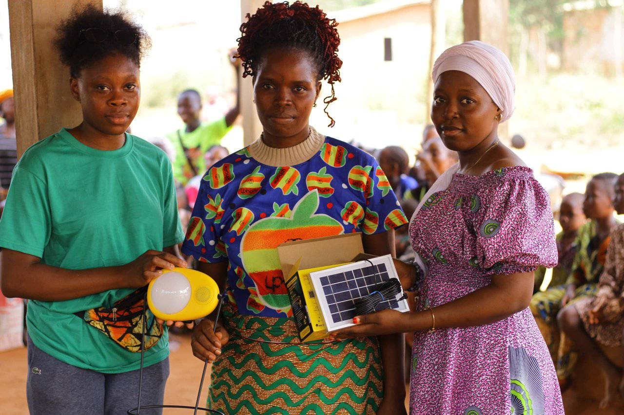 Three Black women pose together for the camera with serious expressions. They are holding pieces of renewable energy technology, including a small solar panel.