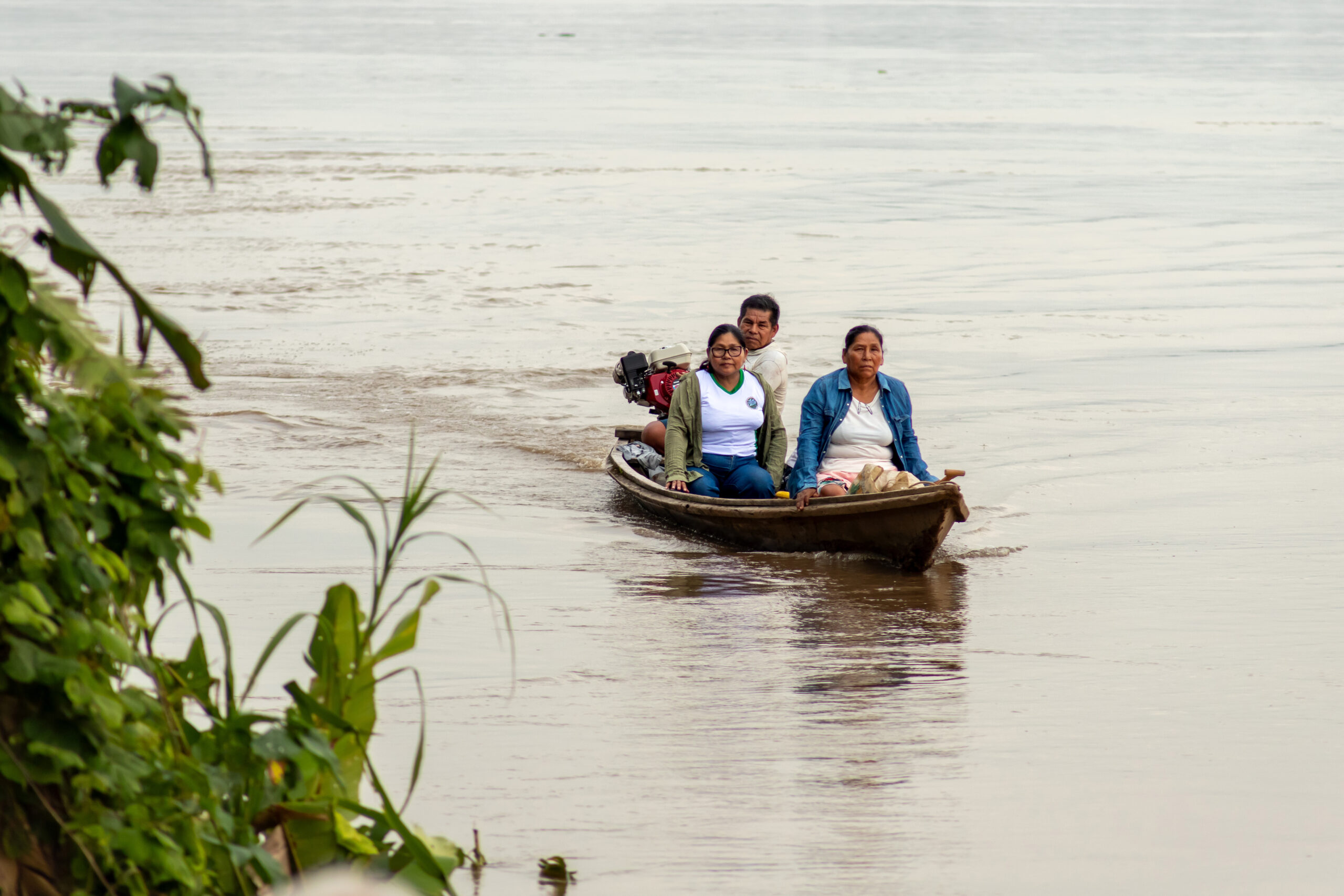 A photo of three Indigenous Peruvian activists sitting together in a small motorboat, a small wake behind it conveying that the boat is moving. Around them is pale river water, and emerging from the bottom corner of the photo are green tree branches.